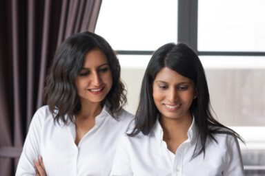 Two smiling female coworkers looking through document in cafe. They are standing with window in background. Document concept. Front view.