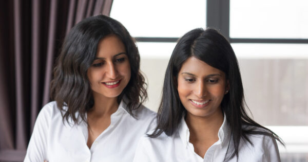 Two smiling female coworkers looking through document in cafe. They are standing with window in background. Document concept. Front view.