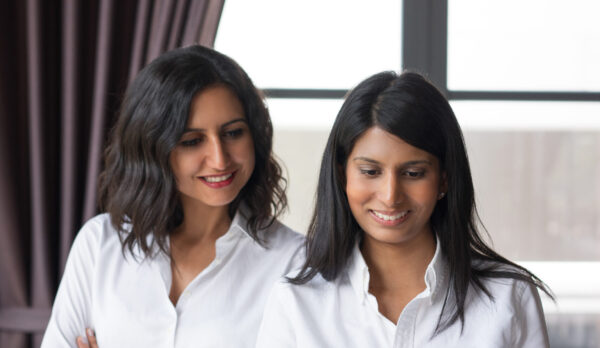 Two smiling female coworkers looking through document in cafe. They are standing with window in background. Document concept. Front view.