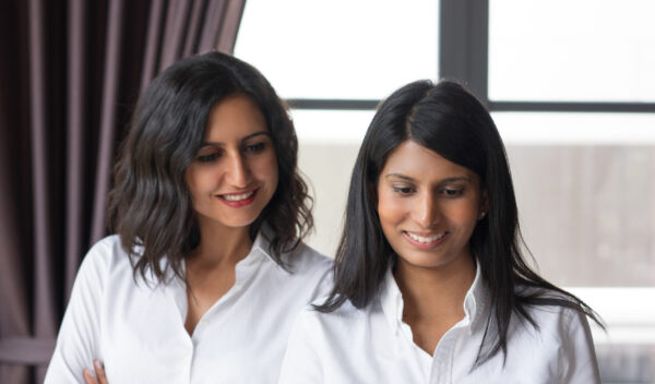 Two smiling female coworkers looking through document in cafe. They are standing with window in background. Document concept. Front view.