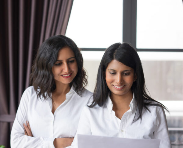 Two smiling female coworkers looking through document in cafe. They are standing with window in background. Document concept. Front view.