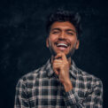 Handsome Indian guy in plaid shirt smiling and holds his hand on chin. Studio photo against a dark textured wall