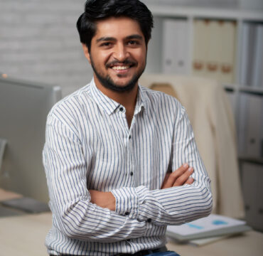 Confident young Indian businessman sitting on edge of his table and looking at camera