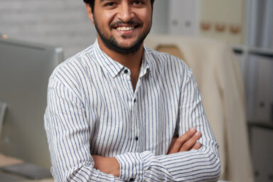 Confident young Indian businessman sitting on edge of his table and looking at camera