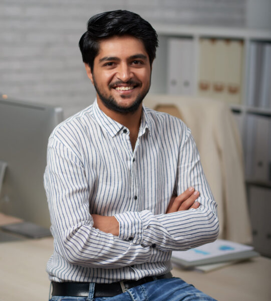 Confident young Indian businessman sitting on edge of his table and looking at camera