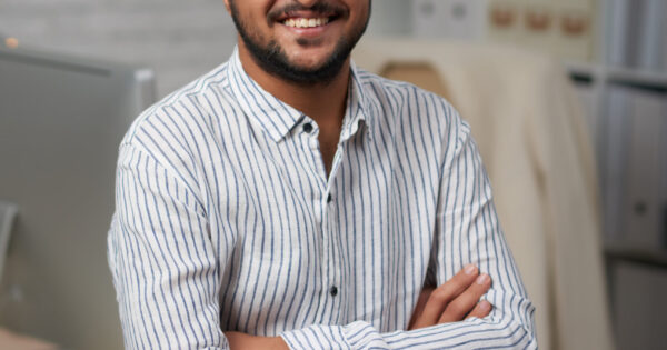 Confident young Indian businessman sitting on edge of his table and looking at camera