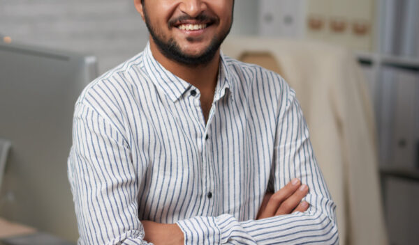 Confident young Indian businessman sitting on edge of his table and looking at camera