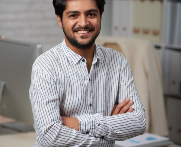 Confident young Indian businessman sitting on edge of his table and looking at camera